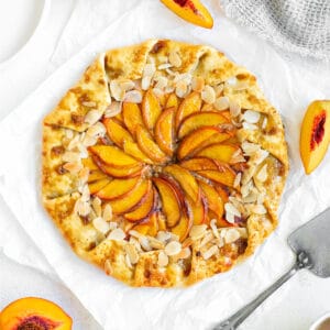 Top view of whole peach galette with almonds on white paper. Peach wedges, silver cake server.