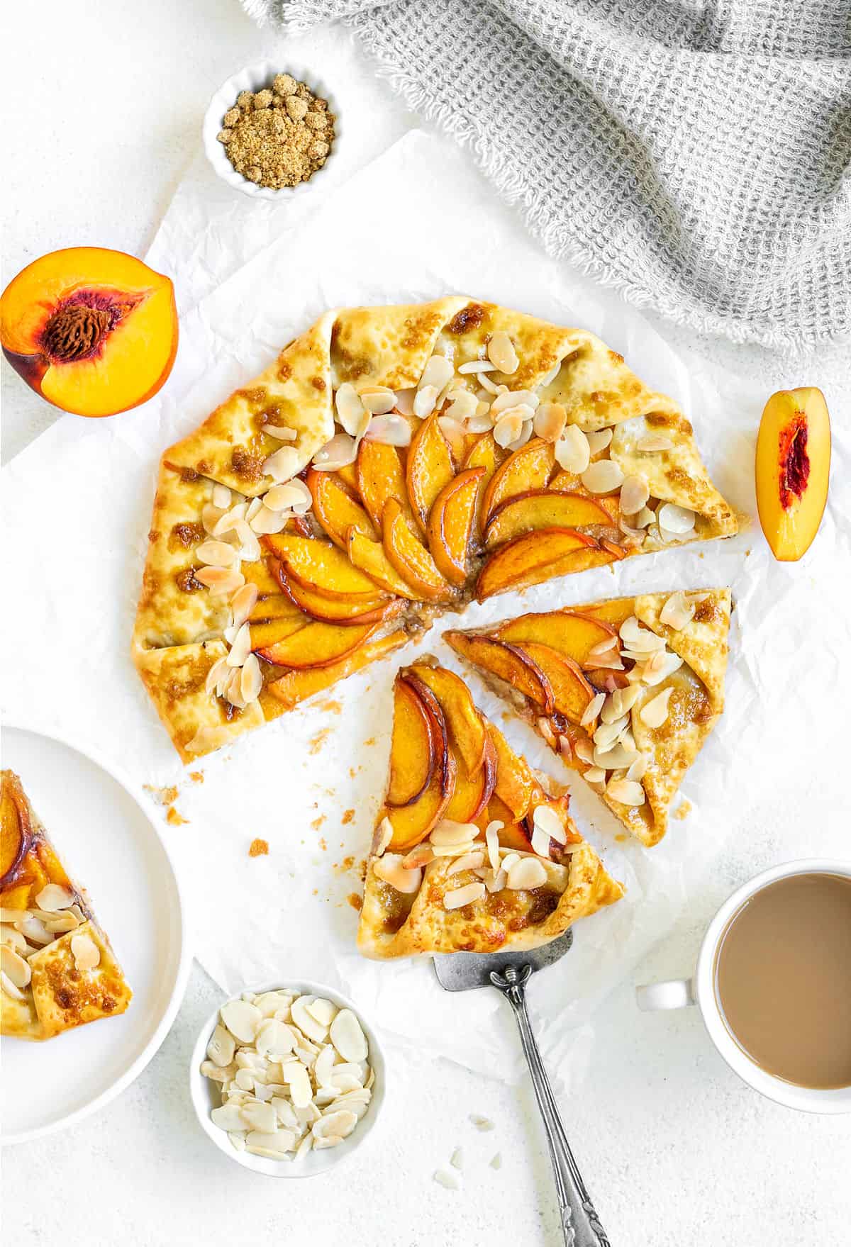 Sliced and whole rustic peach tart on white surface. Cake server, bowls and peach wedges.