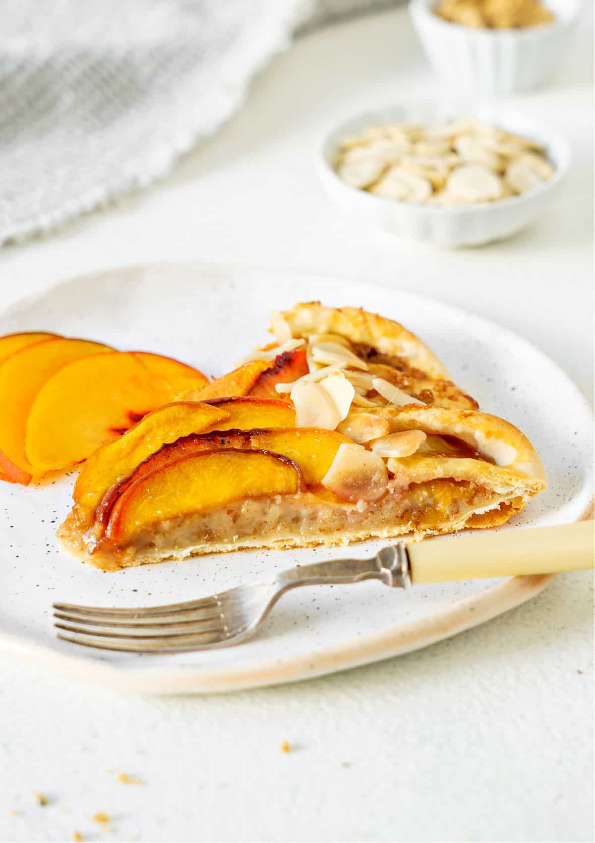 Slice of almond peach galette on a white plate with fork. White background with bowls.