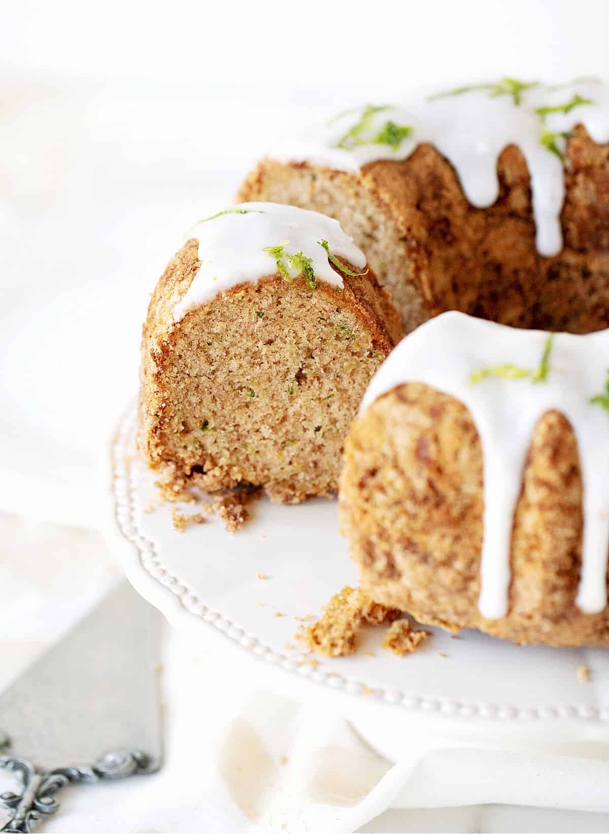 Partial view of glazed bundt cake on white cake stand, silver cake server, white background.