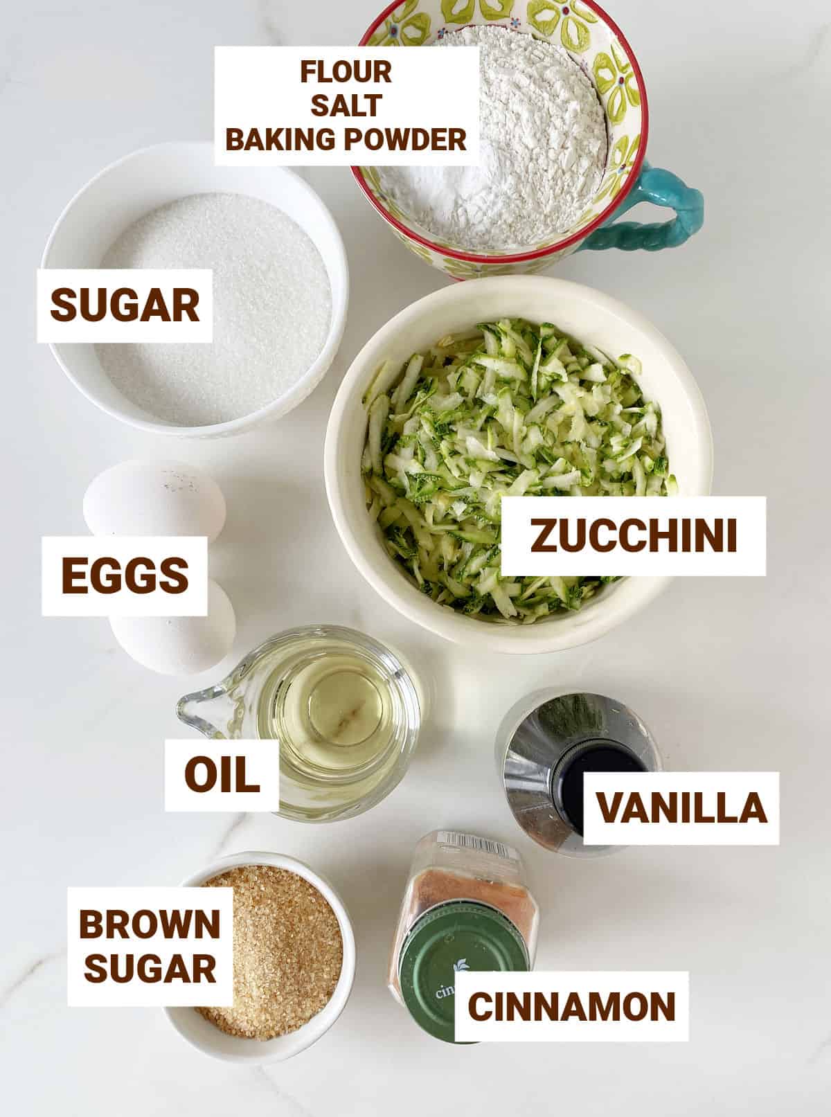 Bowls with zucchini cake ingredients on white surface, including cinnamon, vanilla, oil, brown sugar