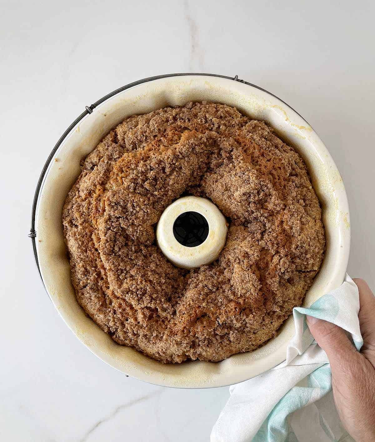 Baked bundt cake in white pan on white surface, hand holding kitchen towel.