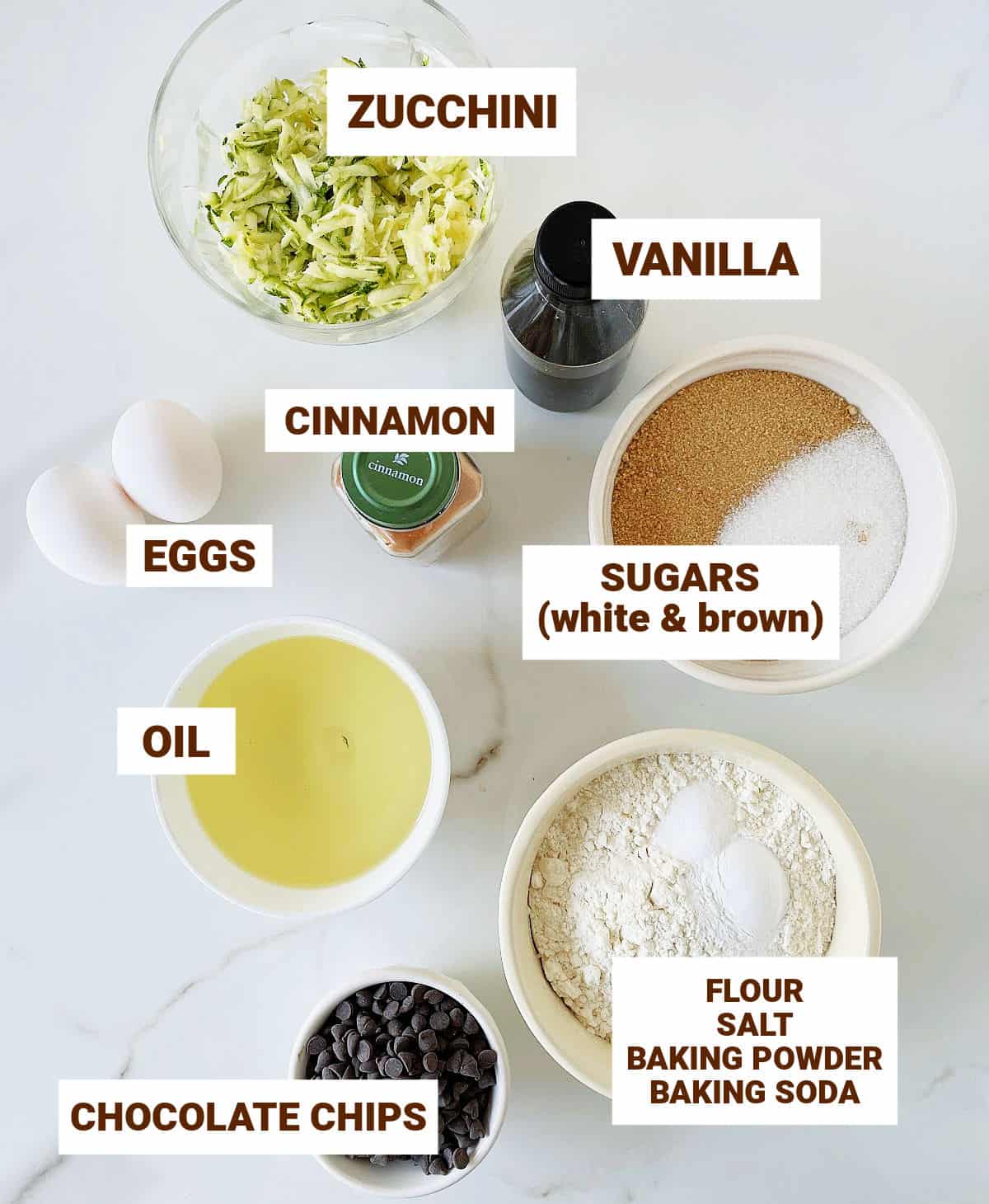 White surface with ingredients for zucchini chocolate chip muffins in bowls including oil, sugars, eggs, vanilla