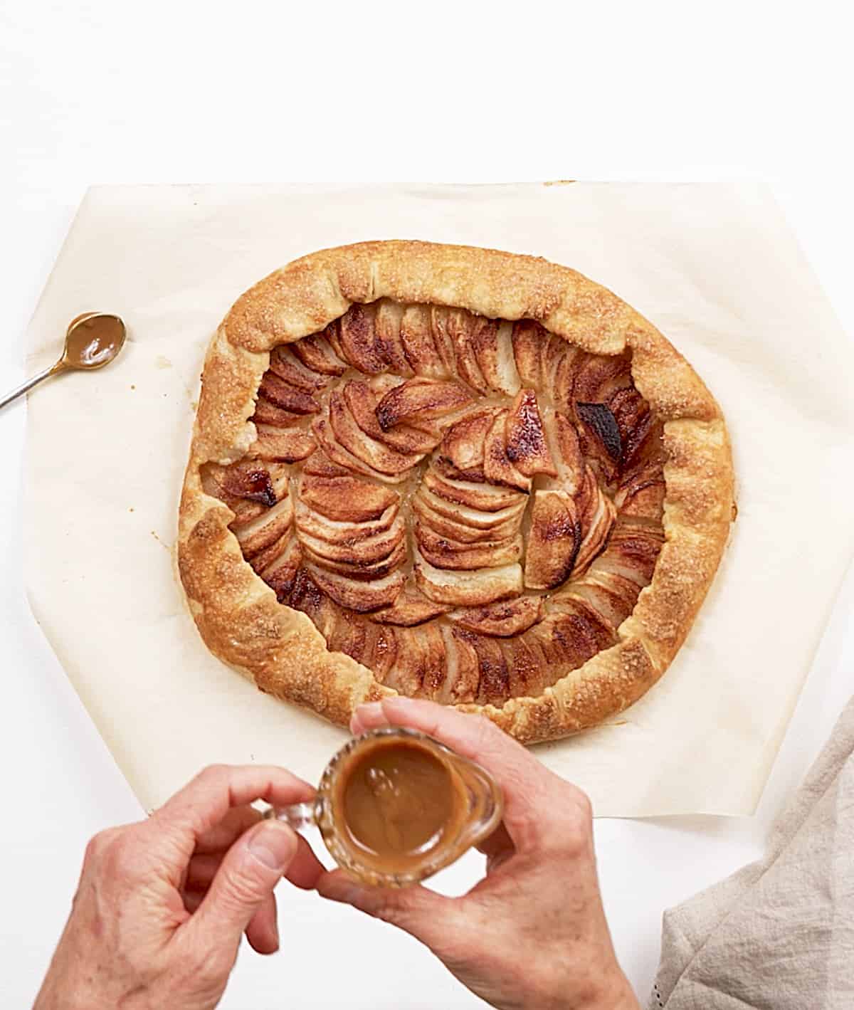 Hands holding glass jar with caramel sauce, an apple galette below on parchment paper