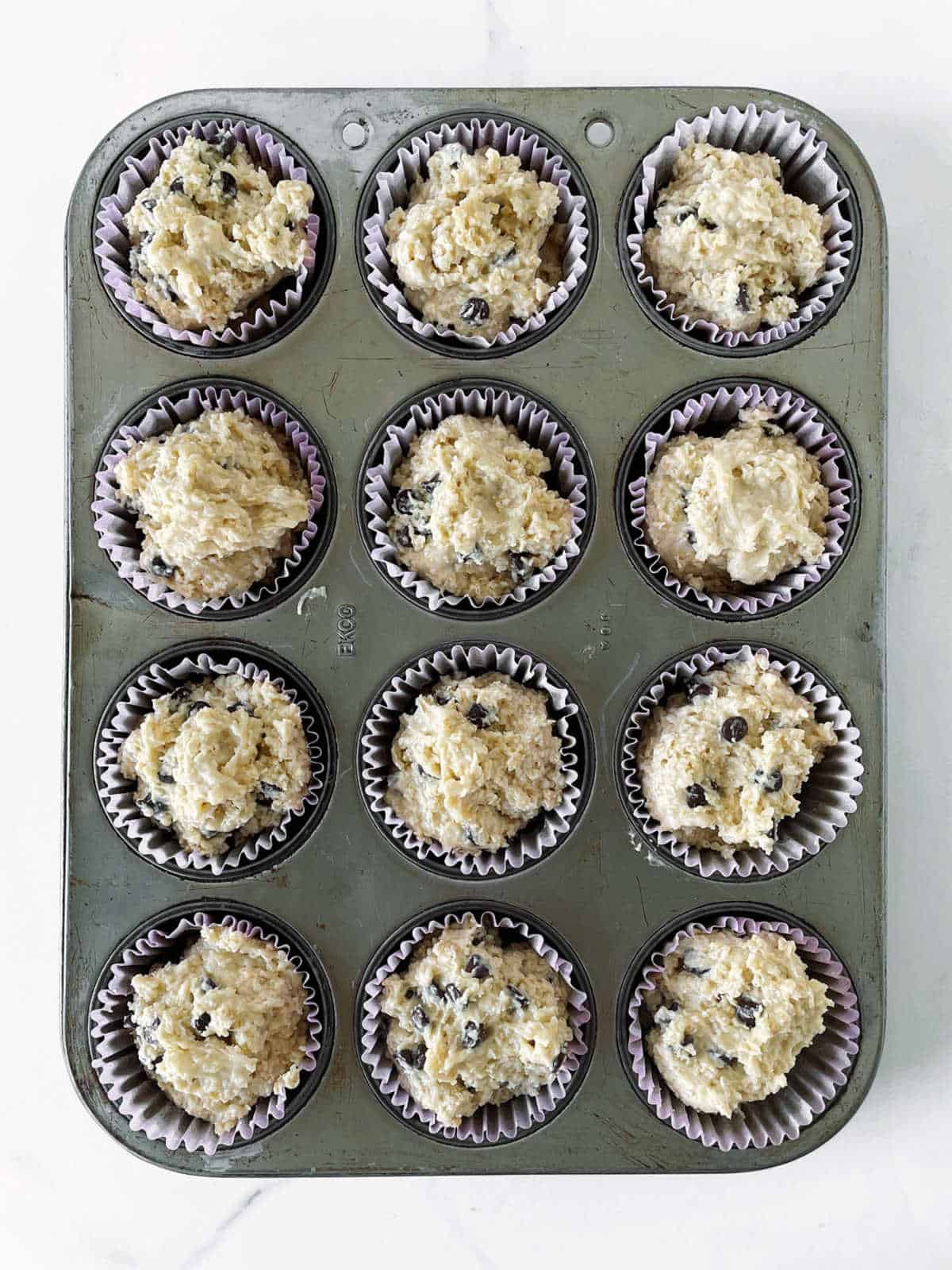 Metal muffin pan with paper liners and oatmeal chocolate chip muffin batter. White surface.