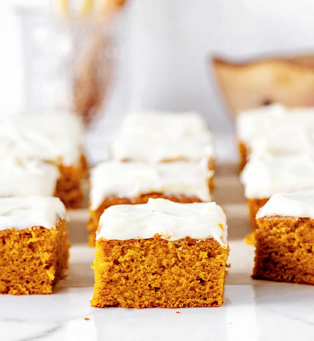 Squares of pumpkin cake with cream cheese frosting on white surface