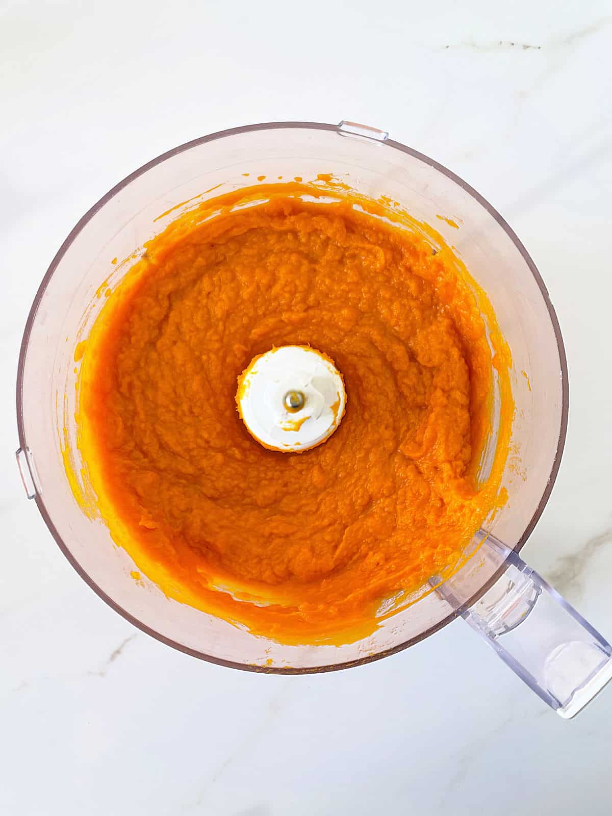 Overview of processed pumpkin puree in the bowl of the processor on white marble