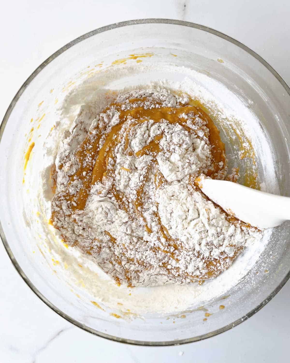 Mixing flour with pumpkin cake batter in a glass bowl with a white spatula. White marble surface.