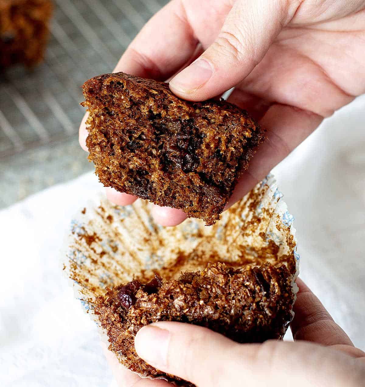 Hands holding a halved raisin bran muffin in a paper liner.
