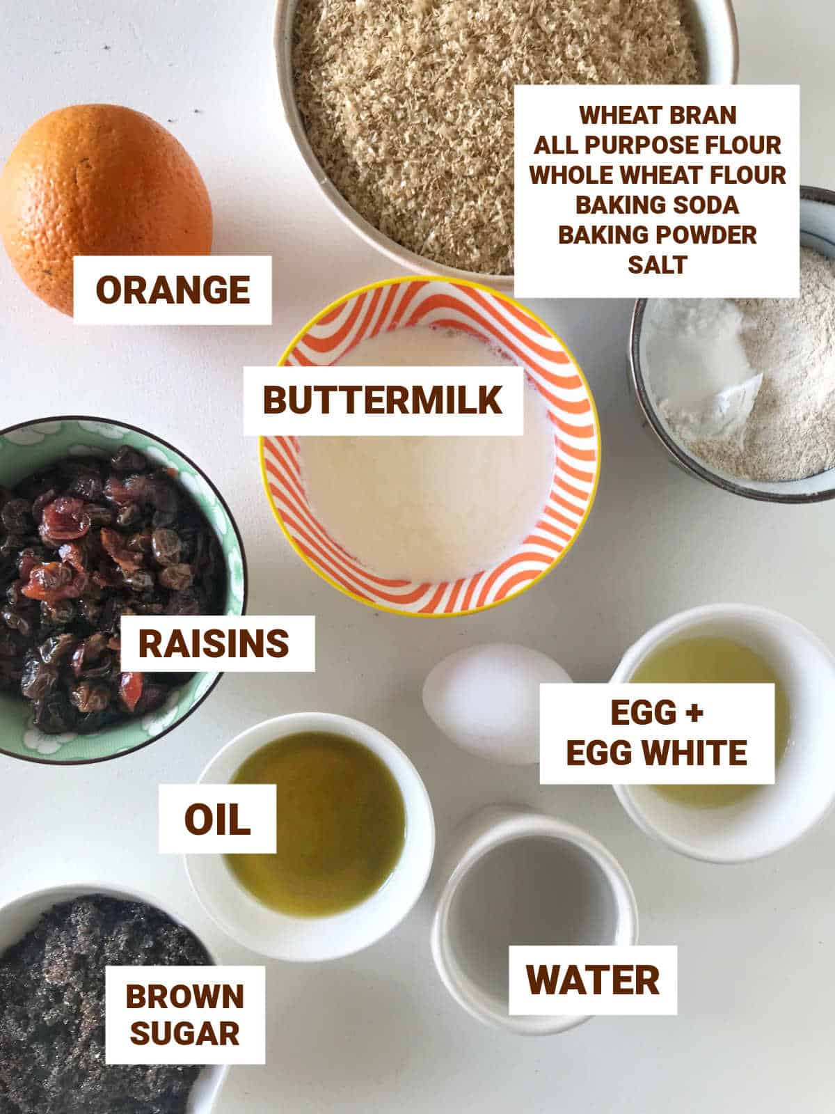 Raisin bran muffin ingredients in bowls on white surface including orange, buttermilk, oil, brown sugar, egg