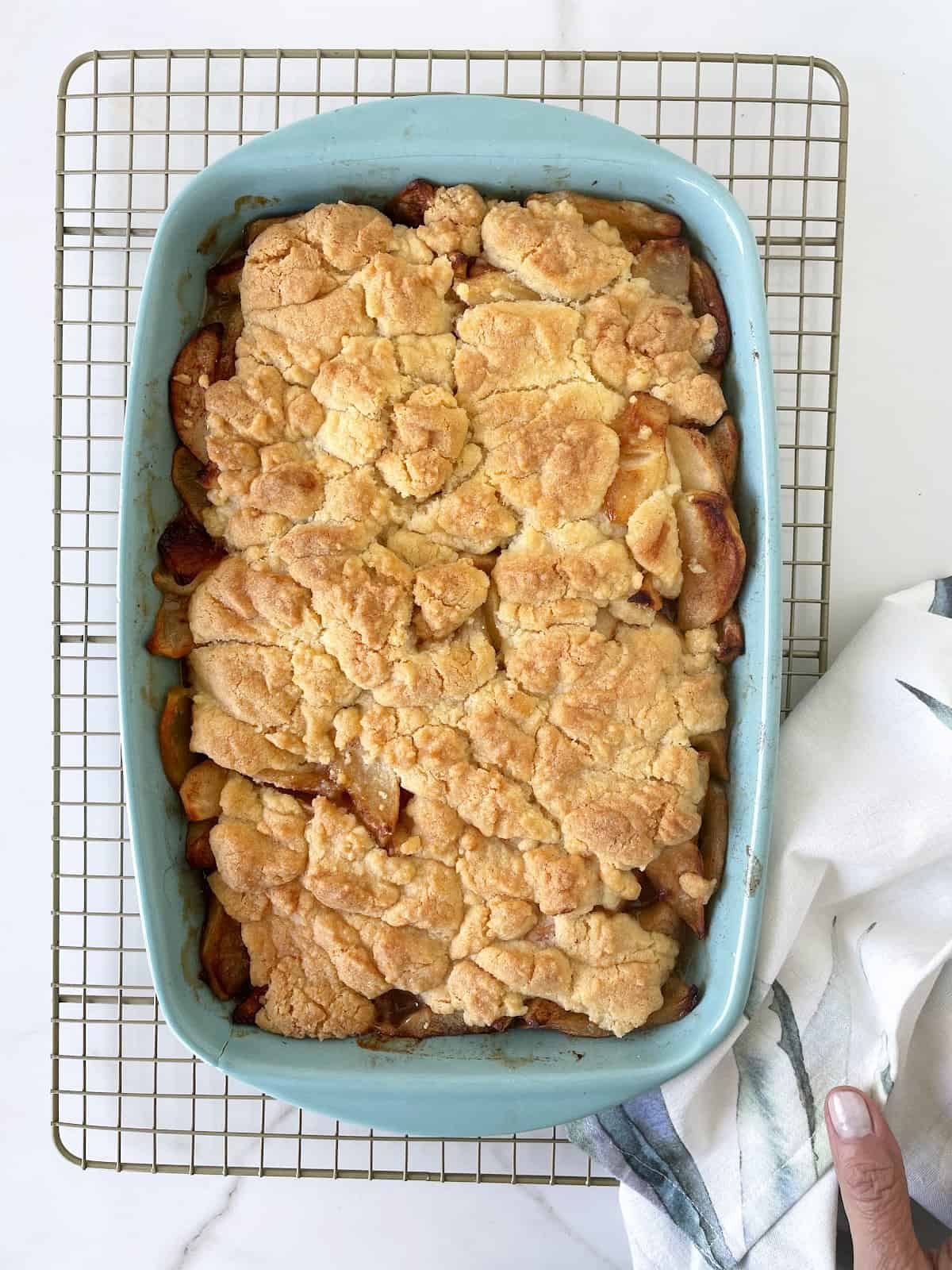 Baked apple dump cake in a blue dish on a wire rack. A kitchen towel. White marble surface.