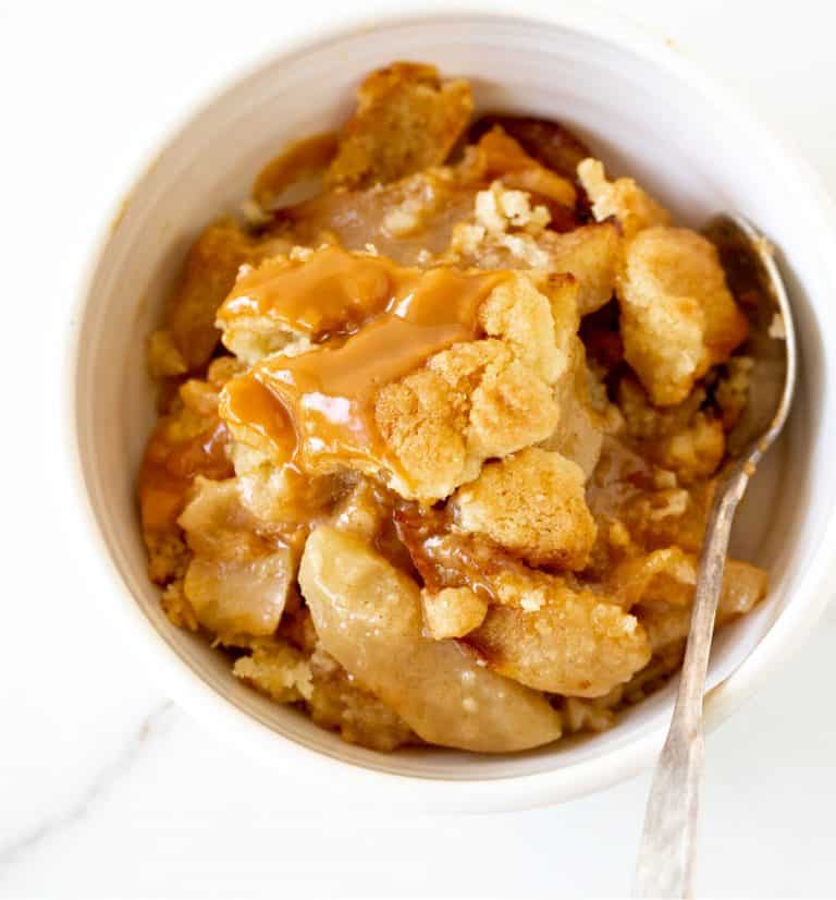 Top view of apple dump cake serving in white bowl with silver spoon over white marble