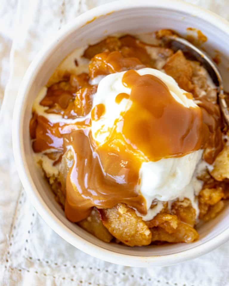 Caramel topped ice cream scoop over apple dump cake in white bowl, white napkin