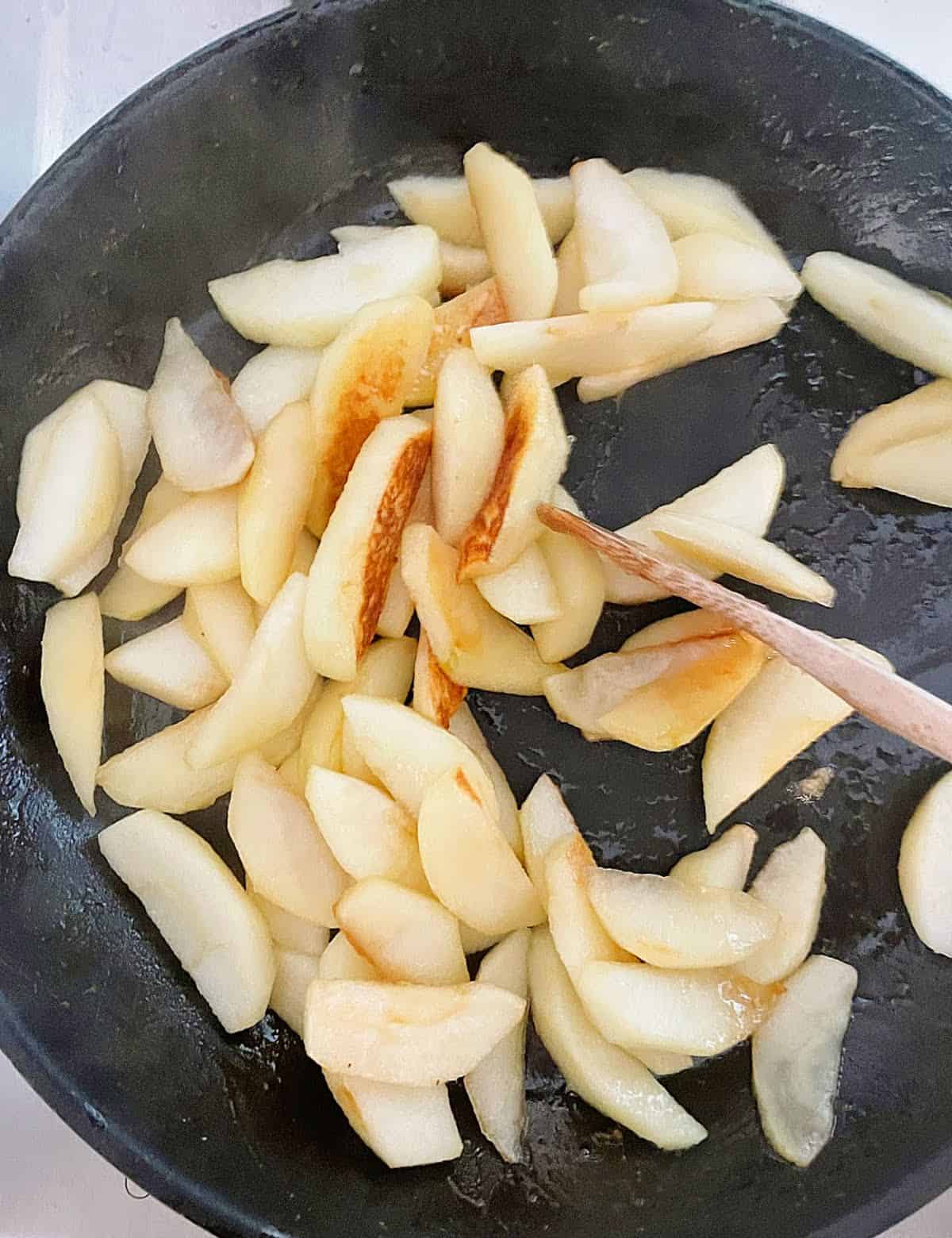 Cooking apple slices in a black skillet with a wooden spoon. 