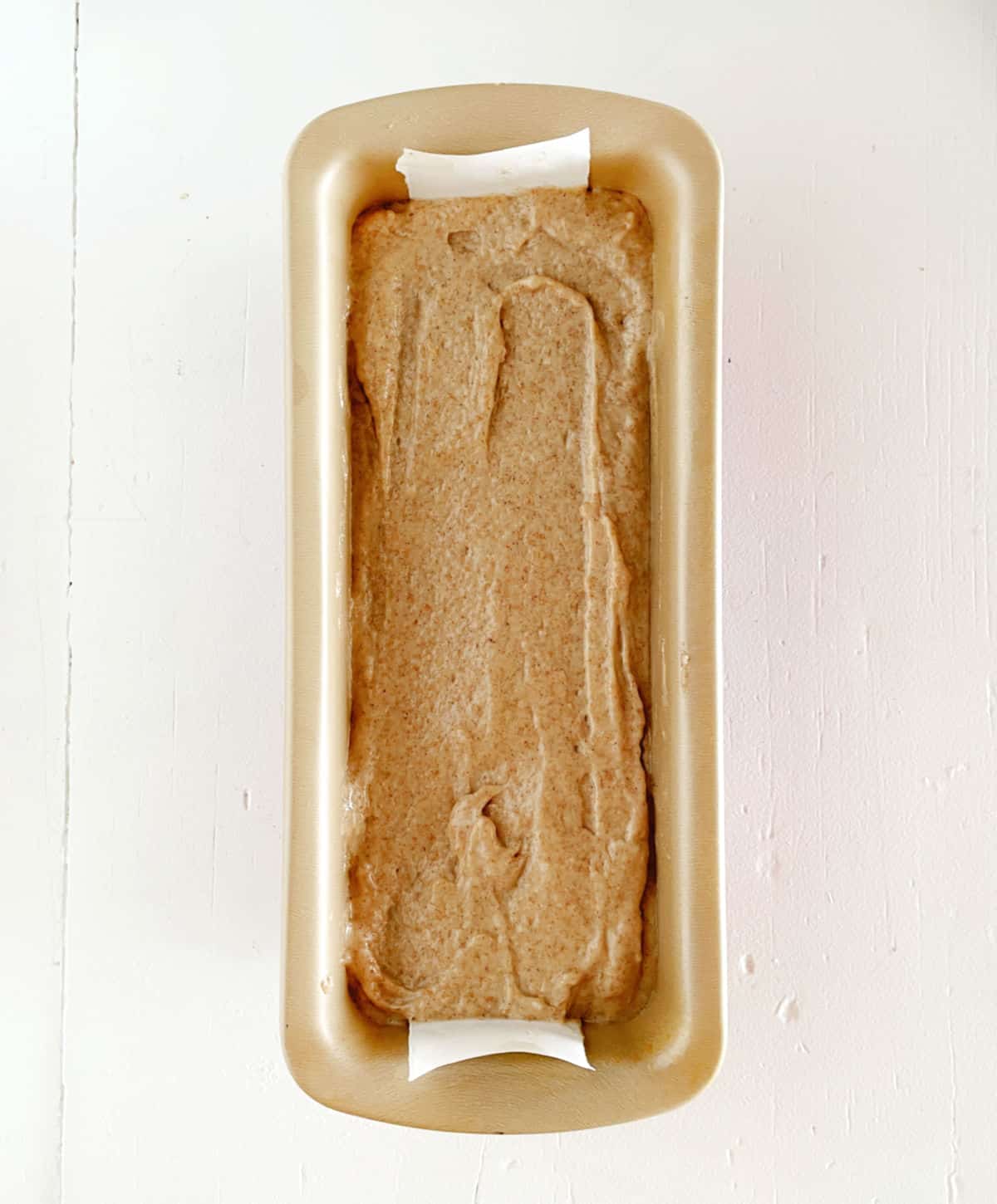 Applesauce bread batter in a gold loaf pan on a white surface. View from above.