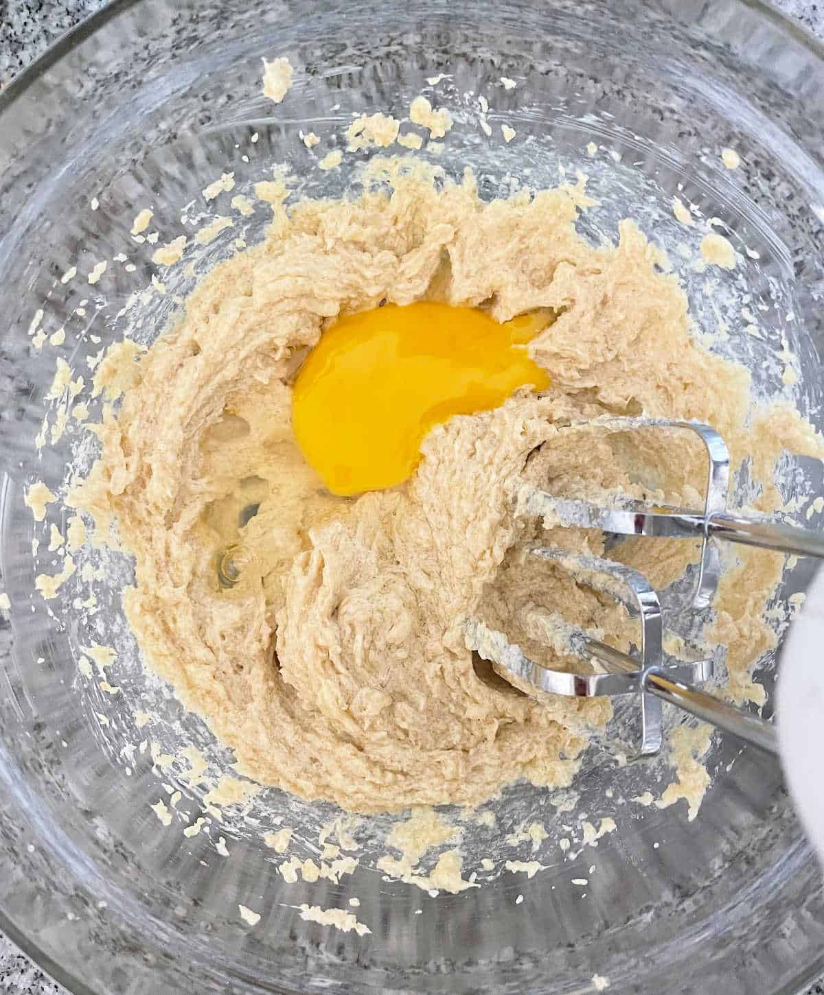 Egg added to butter and sugar mixture in a glass bowl on a grey marble surface. 
