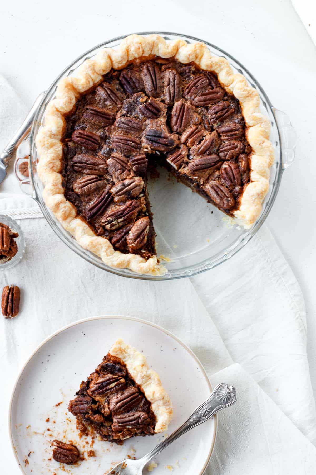 Aerial view of pecan pie in glass dish, slice on a plate with fork, white cloth and surface, loose pecans.