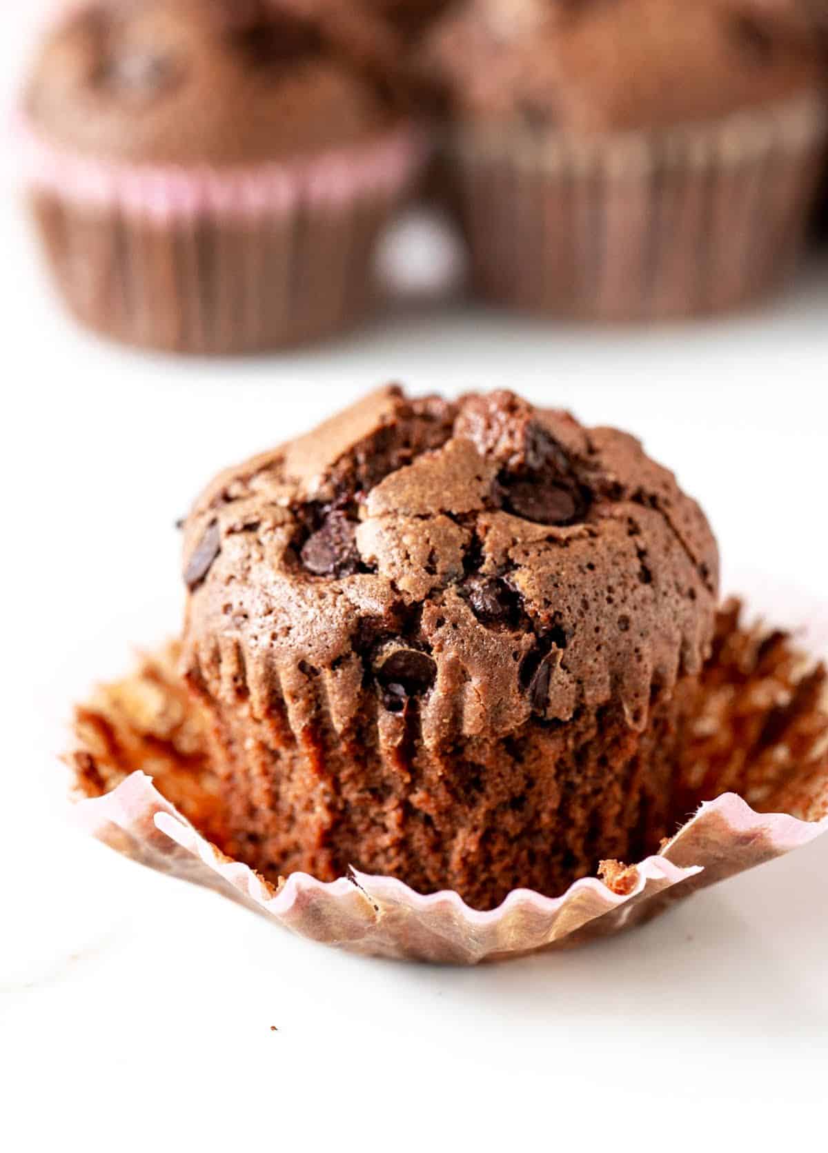 Single chocolate muffin, opened paper liner, white surface, more muffins in background