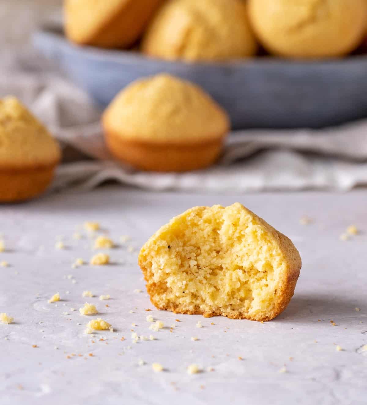 Half cornbread muffin on a light grey surface. More muffins in a grey bowl in the background. 