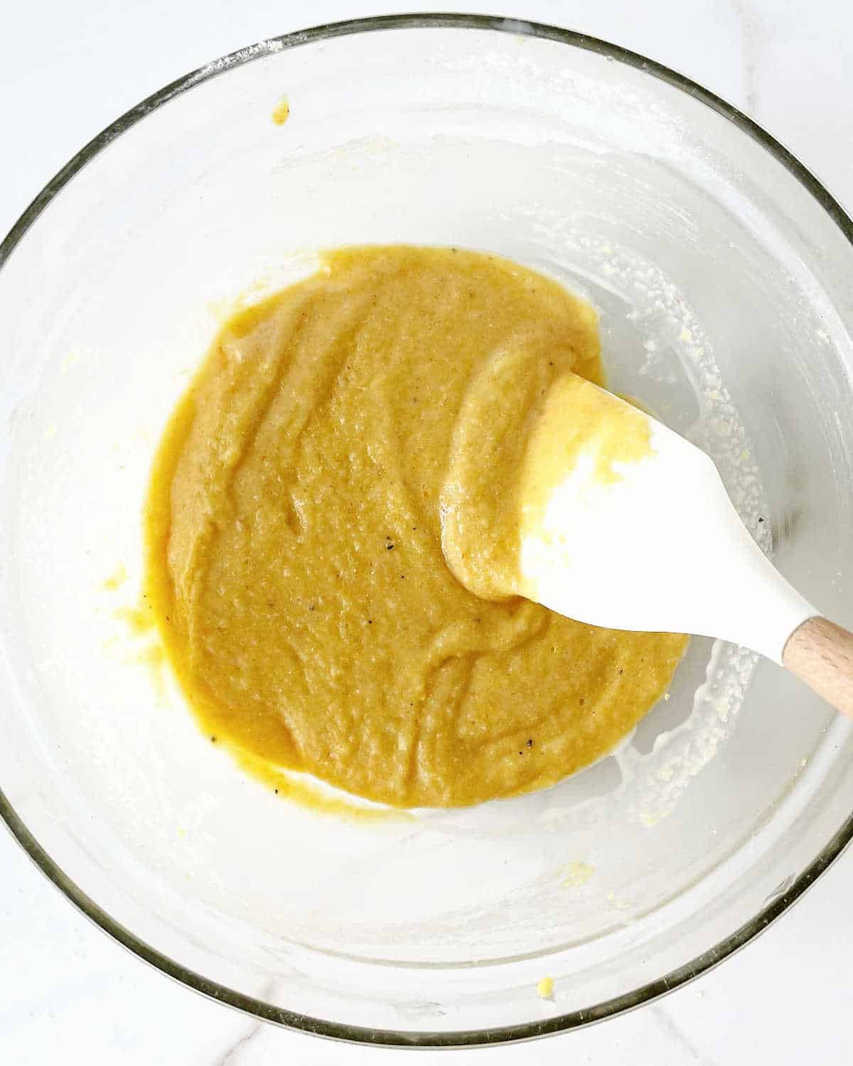 Batter for cornbread muffins in a glass bowl with a white spatula. White marbled surface.