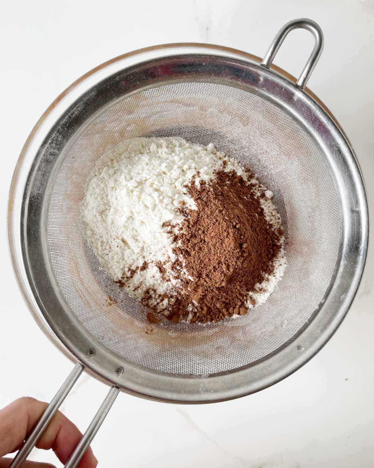 Sifting flour and cocoa powder over a glass bowl on a white surface. 
