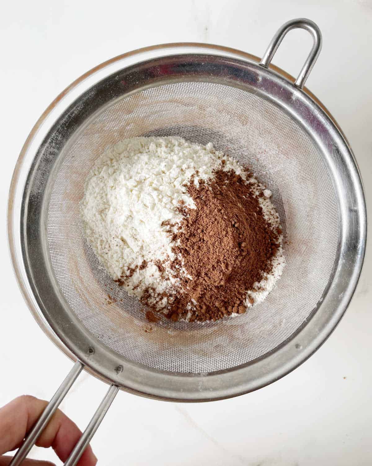 Sifting flour and cocoa powder over a glass bowl on a white surface.