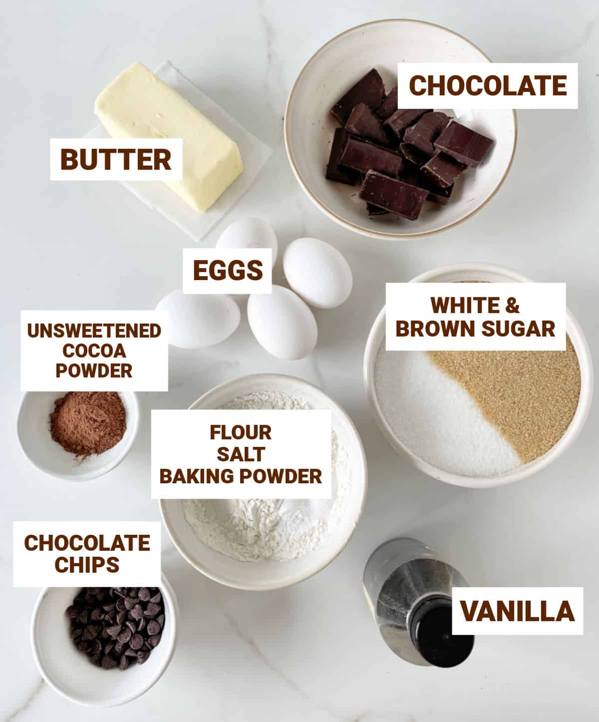 White marble with bowls containing ingredients for chocolate muffins including butter, vanilla, sugar, cocoa powder, chips, eggs.