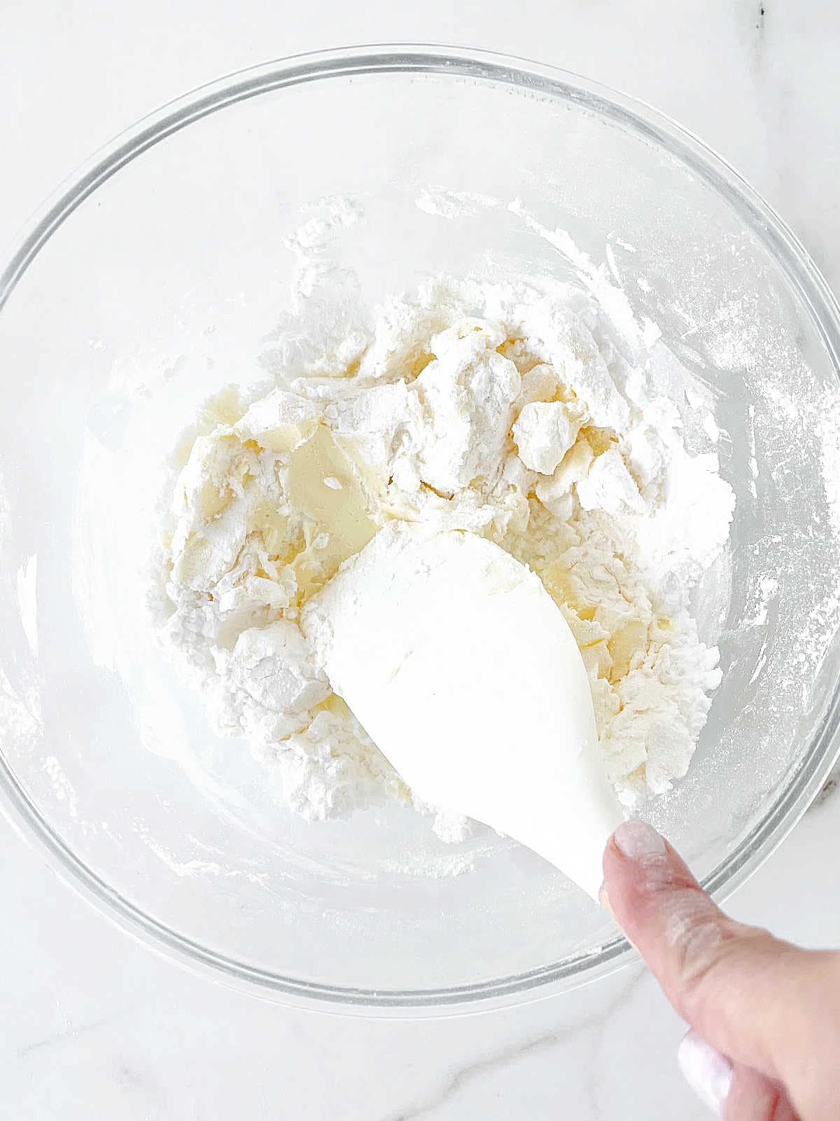Integrating flour into cookie dough mixture in a glass bowl on a white surface with white spatula.