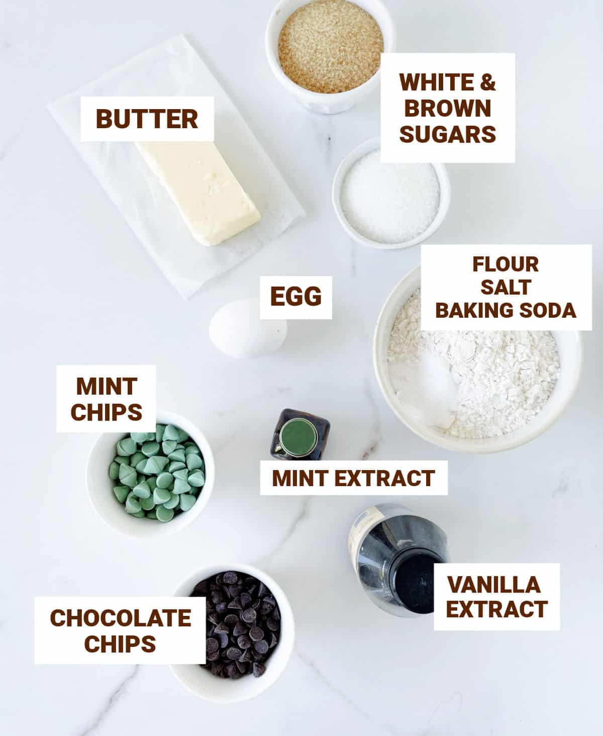 White marble surface with bowls containing ingredients for mint chocolate chip cookies including sugars, mint chips, butter, flour, vanilla.