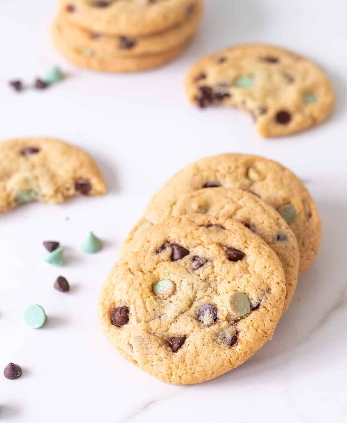 Several whole and bitten mint chocolate chip cookies on white marble with extra chips around.