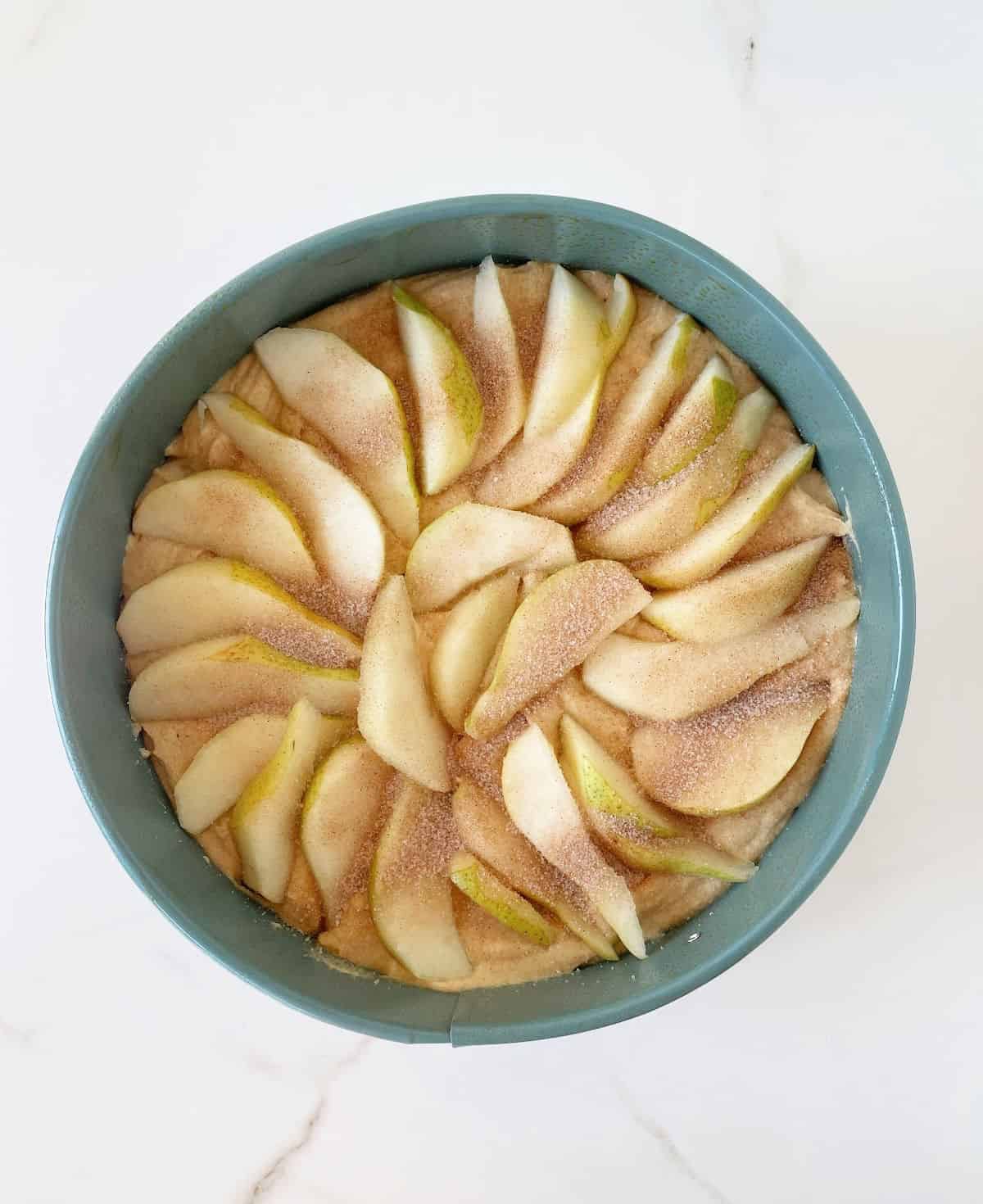 Pear slices with cinnamon sugar on cake batter on a teal round pan on white marble surface. 