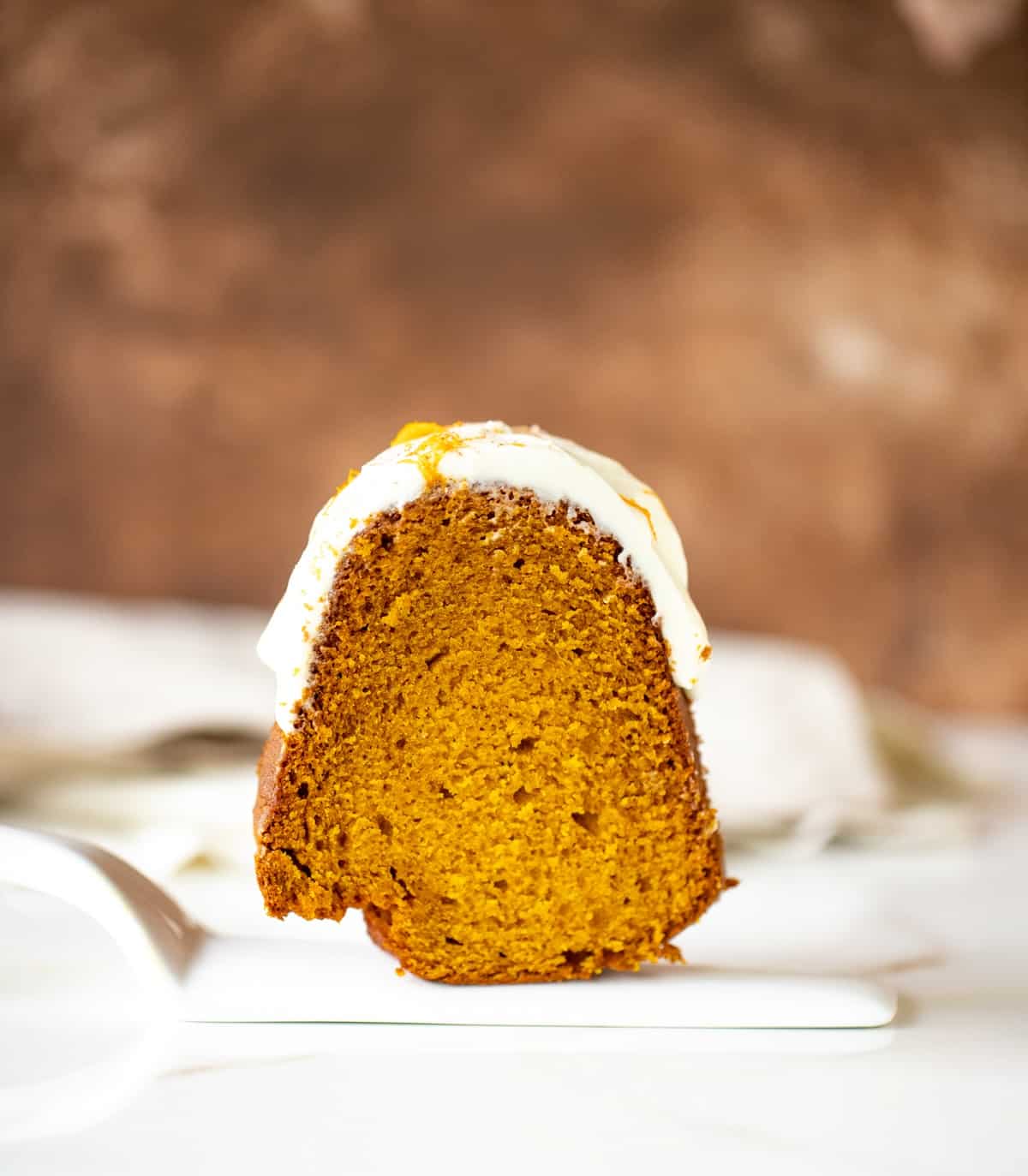 Single slice of frosted pumpkin bundt cake on white cake server, white and brown background