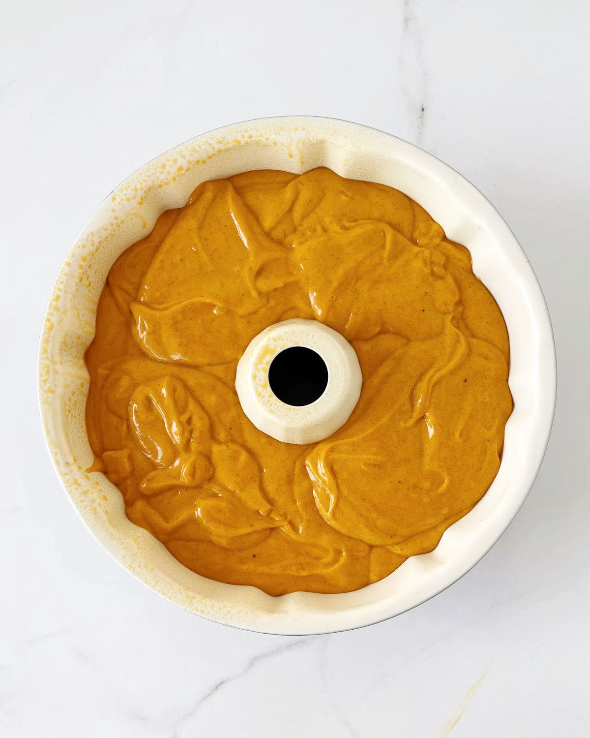 Pumpkin cake batter in a bundt pan on a white marbled surface.