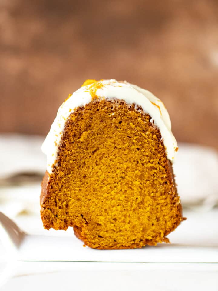 Single frosted slice of pumpkin bundt cake on a white ceramic cake server. Brown background.