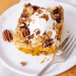 A serving of cream topped pumpkin crumb cake on white plate, silver fork, orange beige surface