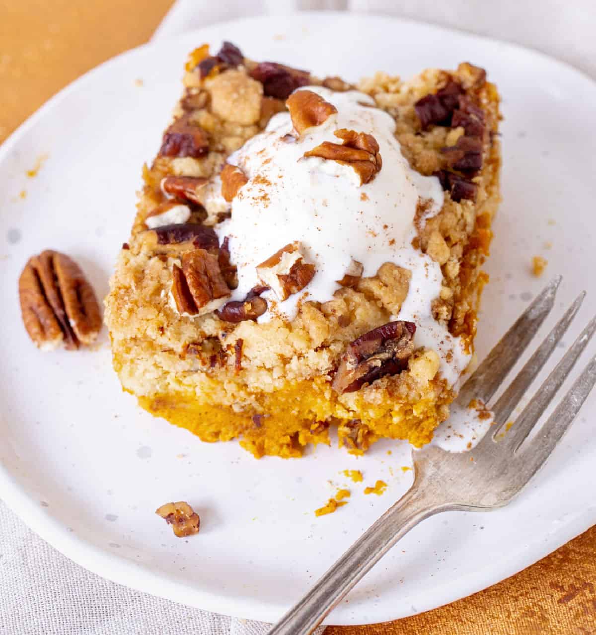 A serving of cream topped pumpkin crumb cake on white plate, silver fork, orange beige surface