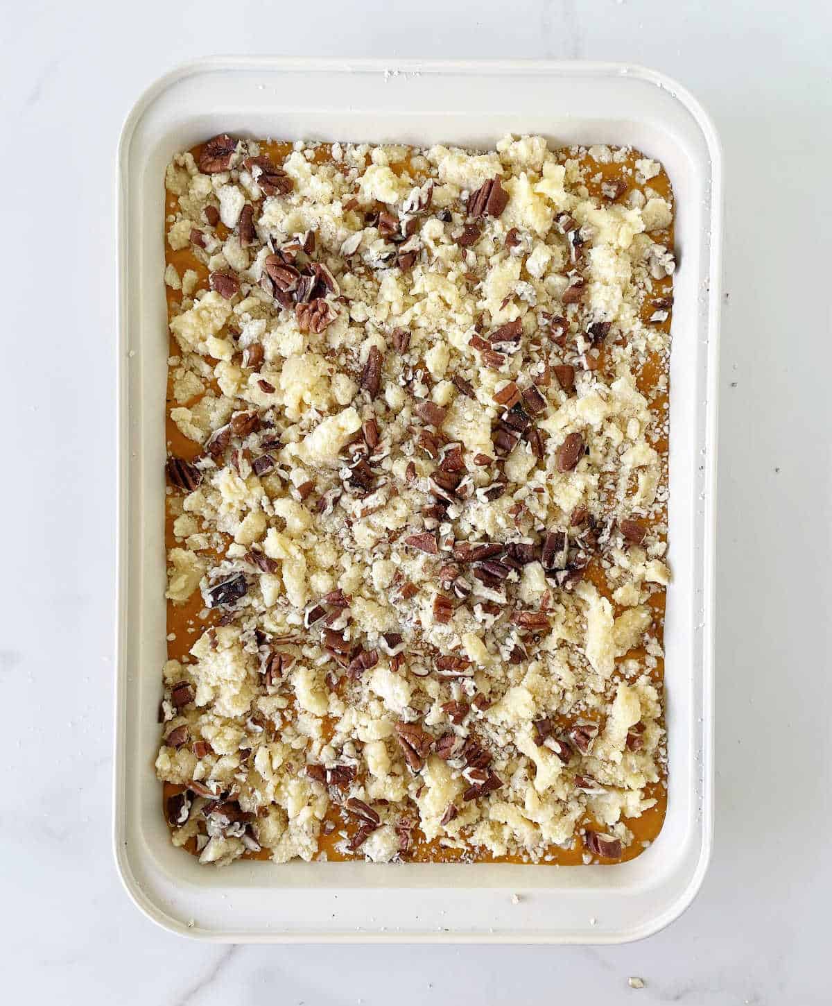 Ready to be baked pumpkin dump cake with pecans on a white rectangular pan on a white marble surface.