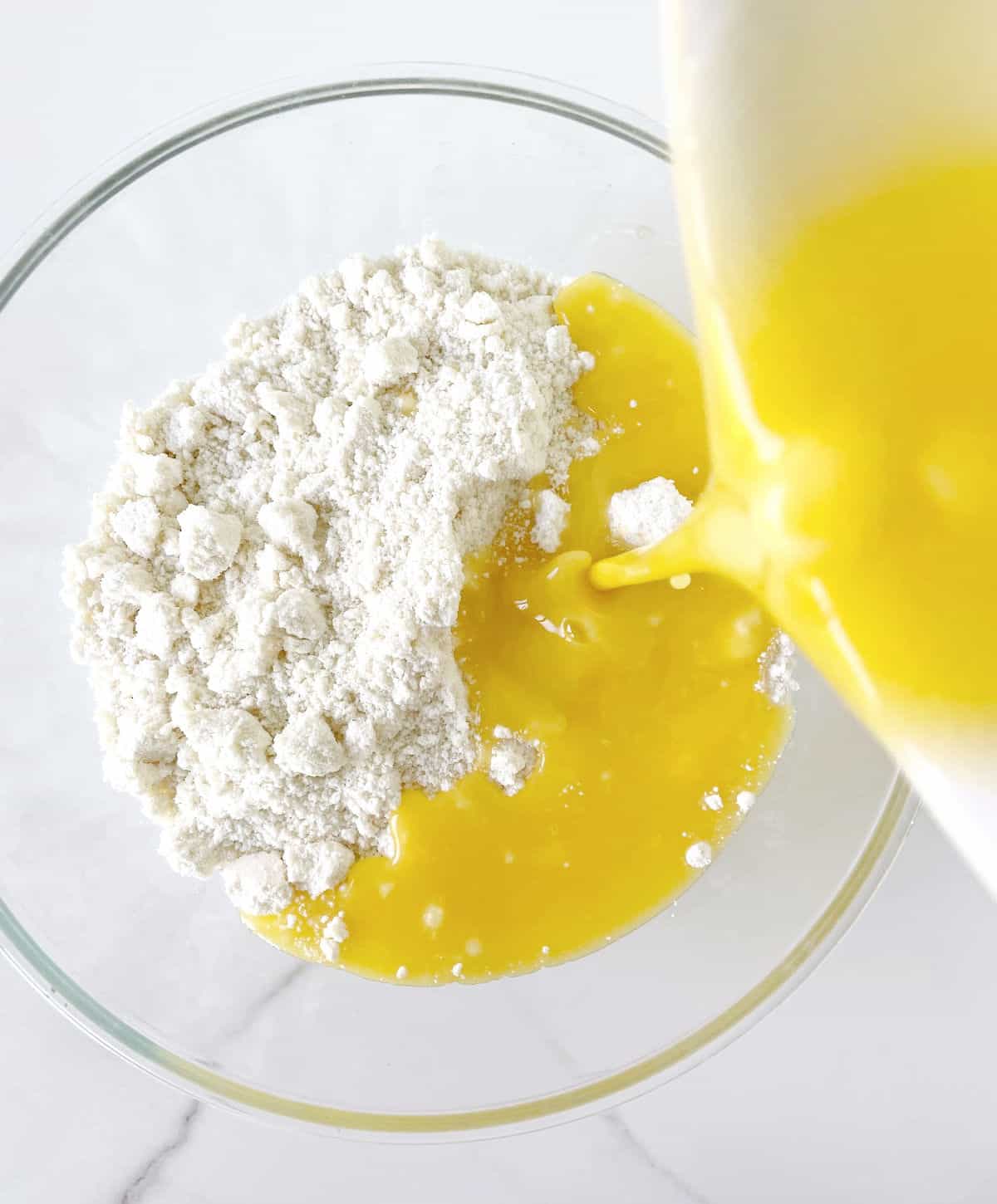 Adding melted butter to white cake mix in a glass bowl on a white marble surface.