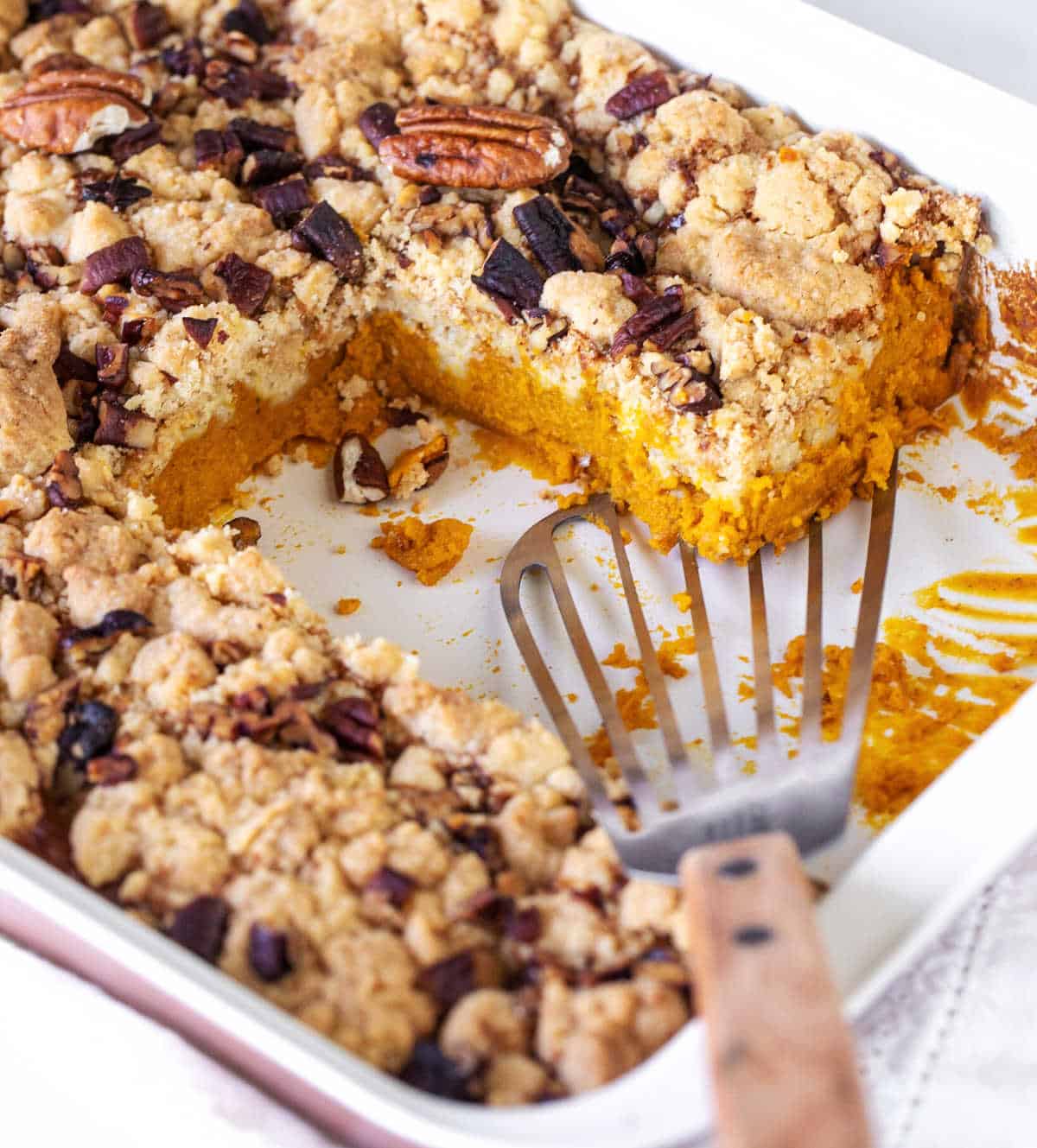 Close of baking dish with pumpkin crumb cake, portions missing, a metal serving spatula.