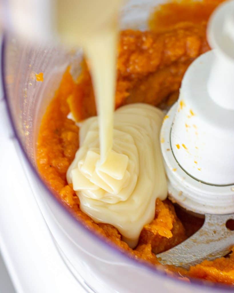 Condensed milk being poured into the jar of a food processor with pumpkin puree.