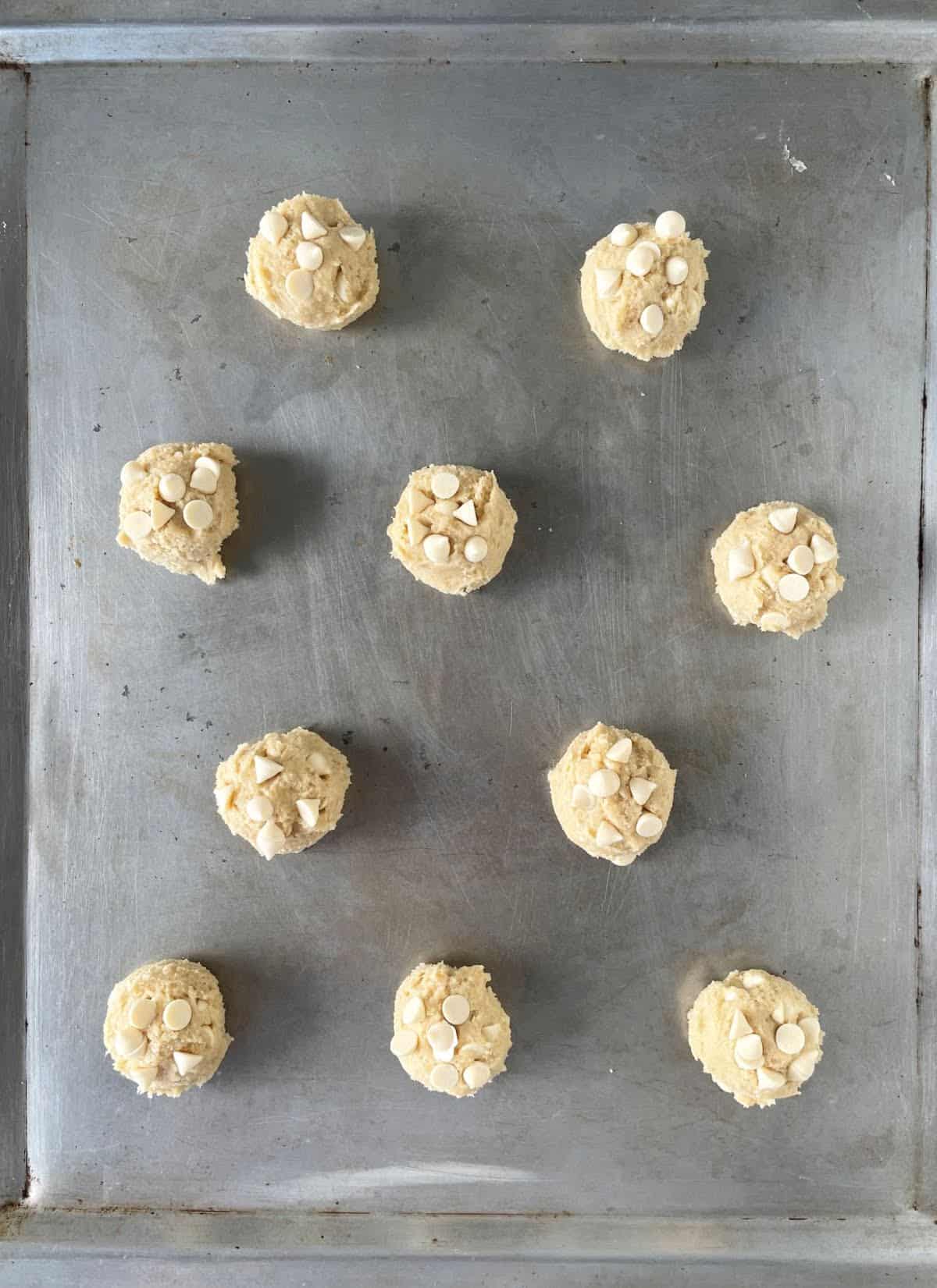 Baking metal sheet with scoops of unbaked white chocolate cookies.