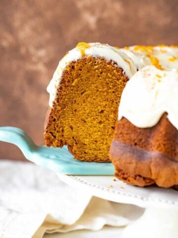 A slice of frosted pumpkin bundt cake on blue cake server, brown background, cake on stand
