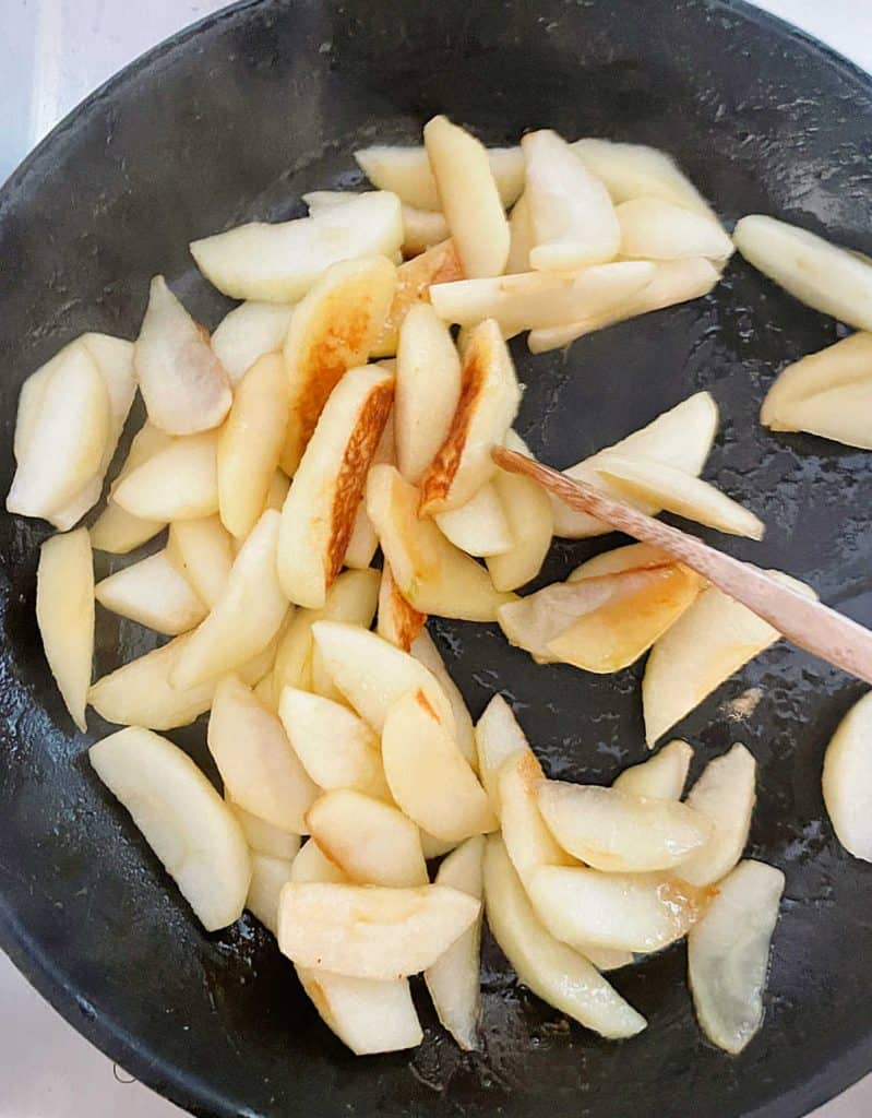 Apple slices being cooked in black skillet, a wooden spoon