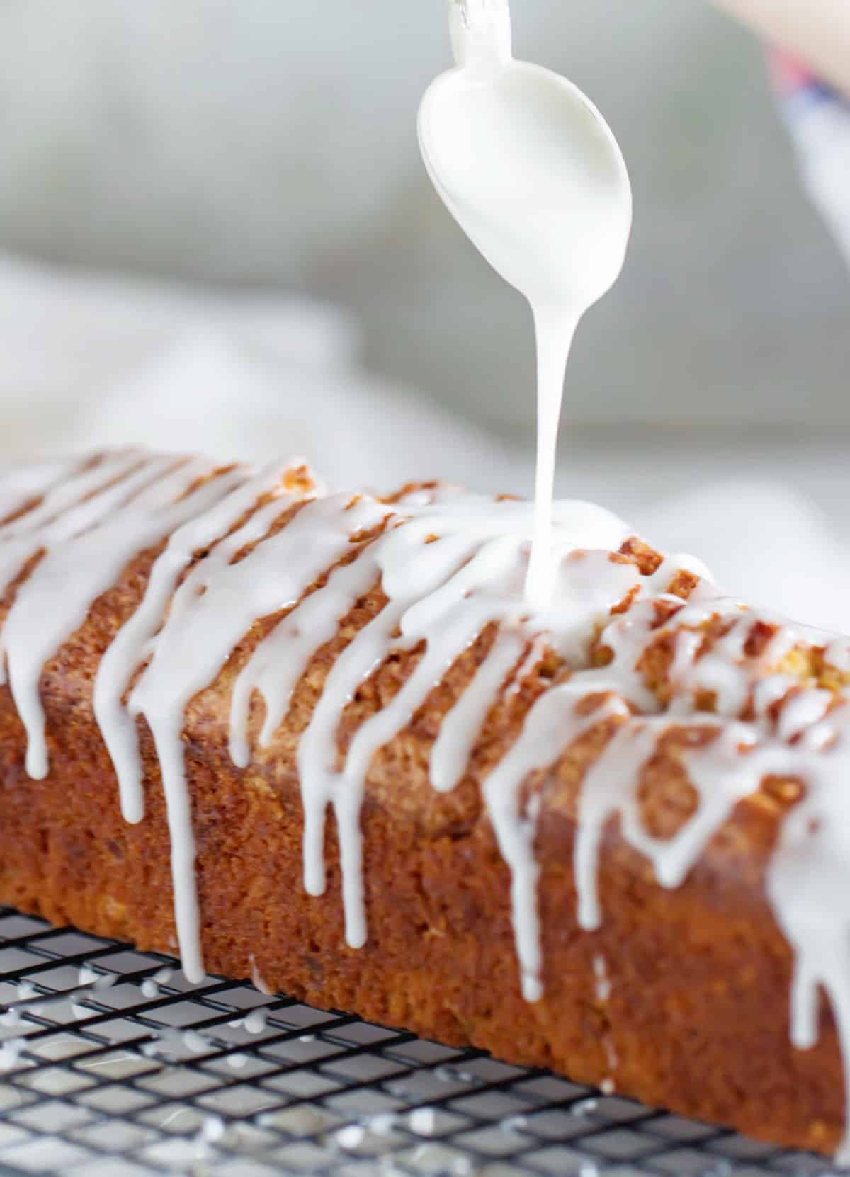 Pouring white glaze from spoon on top of a loaf cake on a wire rack.