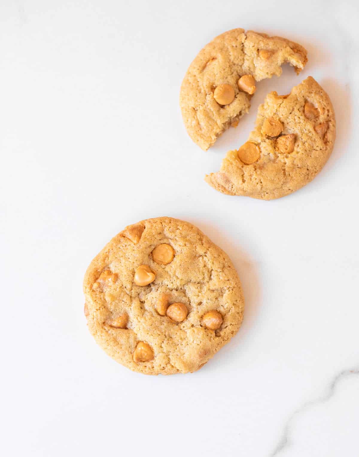 One whole and one halved butterscotch chip cookies on white marble surface. 