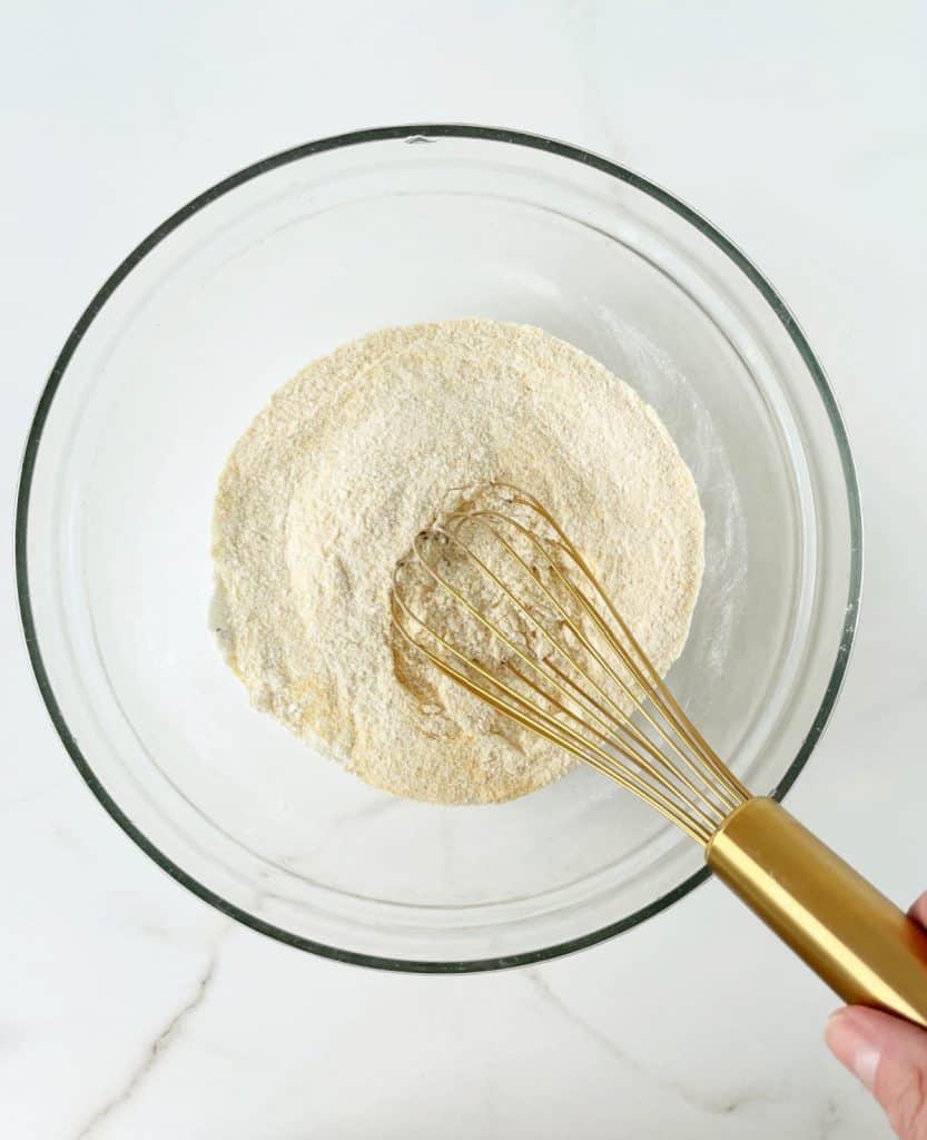 Cornbread muffin dry ingredients stirred in a glass bowl. Golden whisk. White surface.