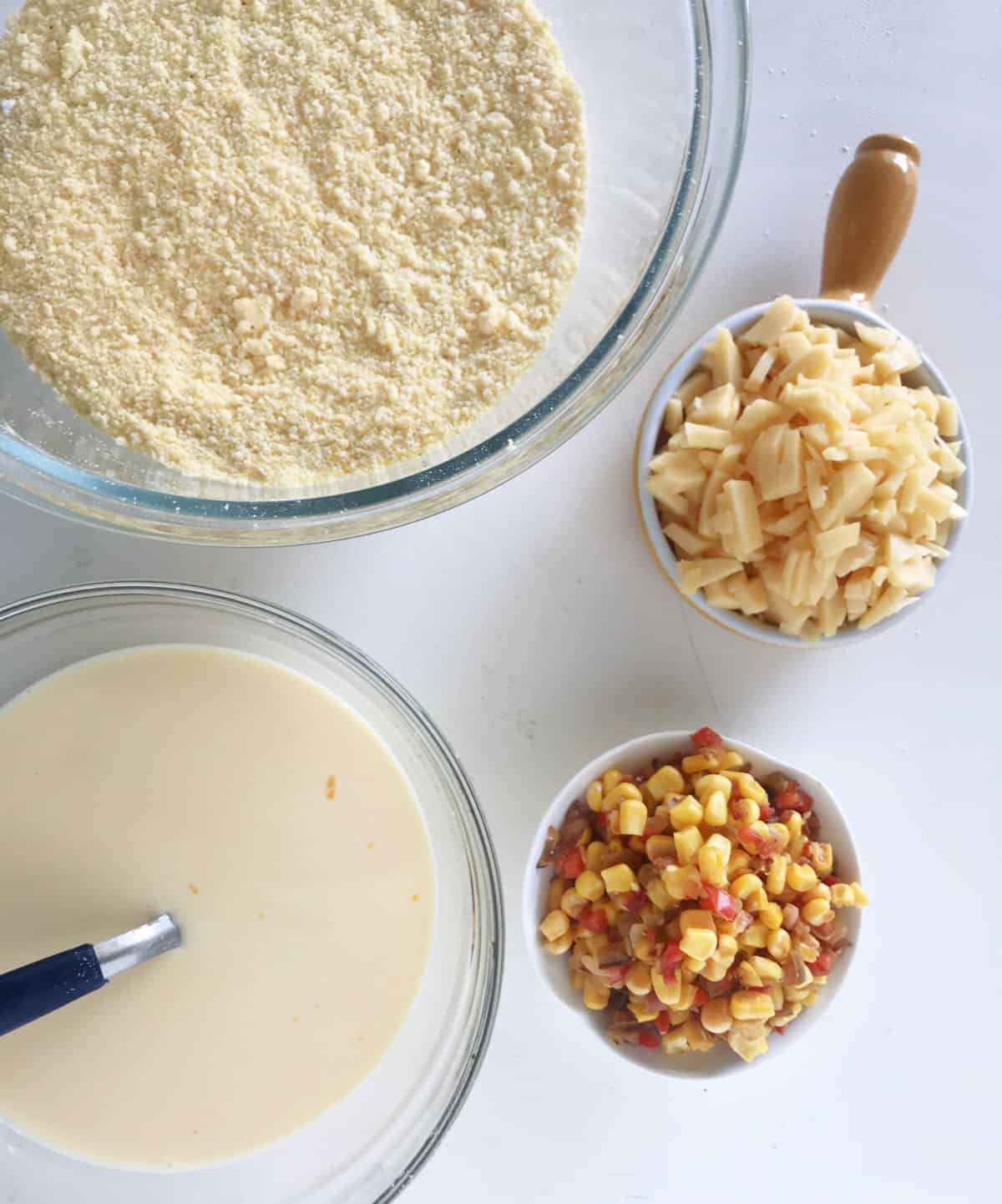 White surface with bowls containing cheese, corn, cornmeal and wet ingredients