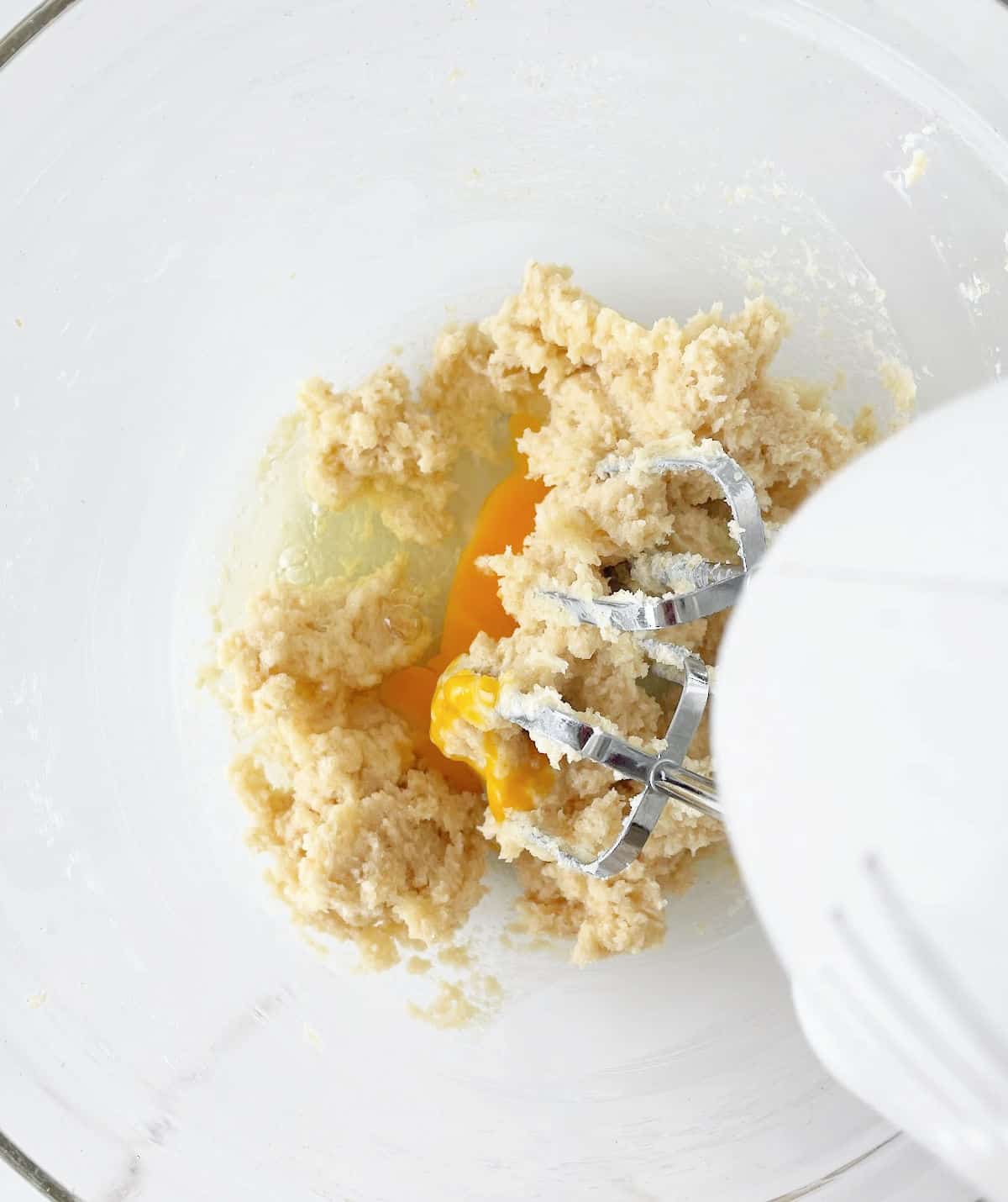 Eggs added to cookie dough mixture in a glass bowl with an electric mixer. White marble surface.