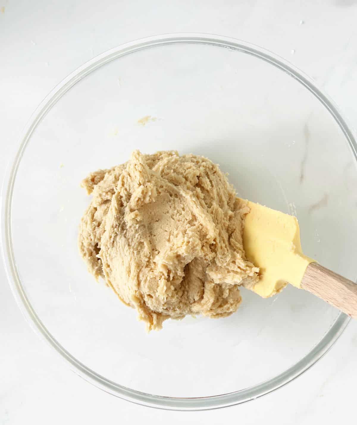 Cookie dough in a glass bowl with a yellow spatula. White marble surface.