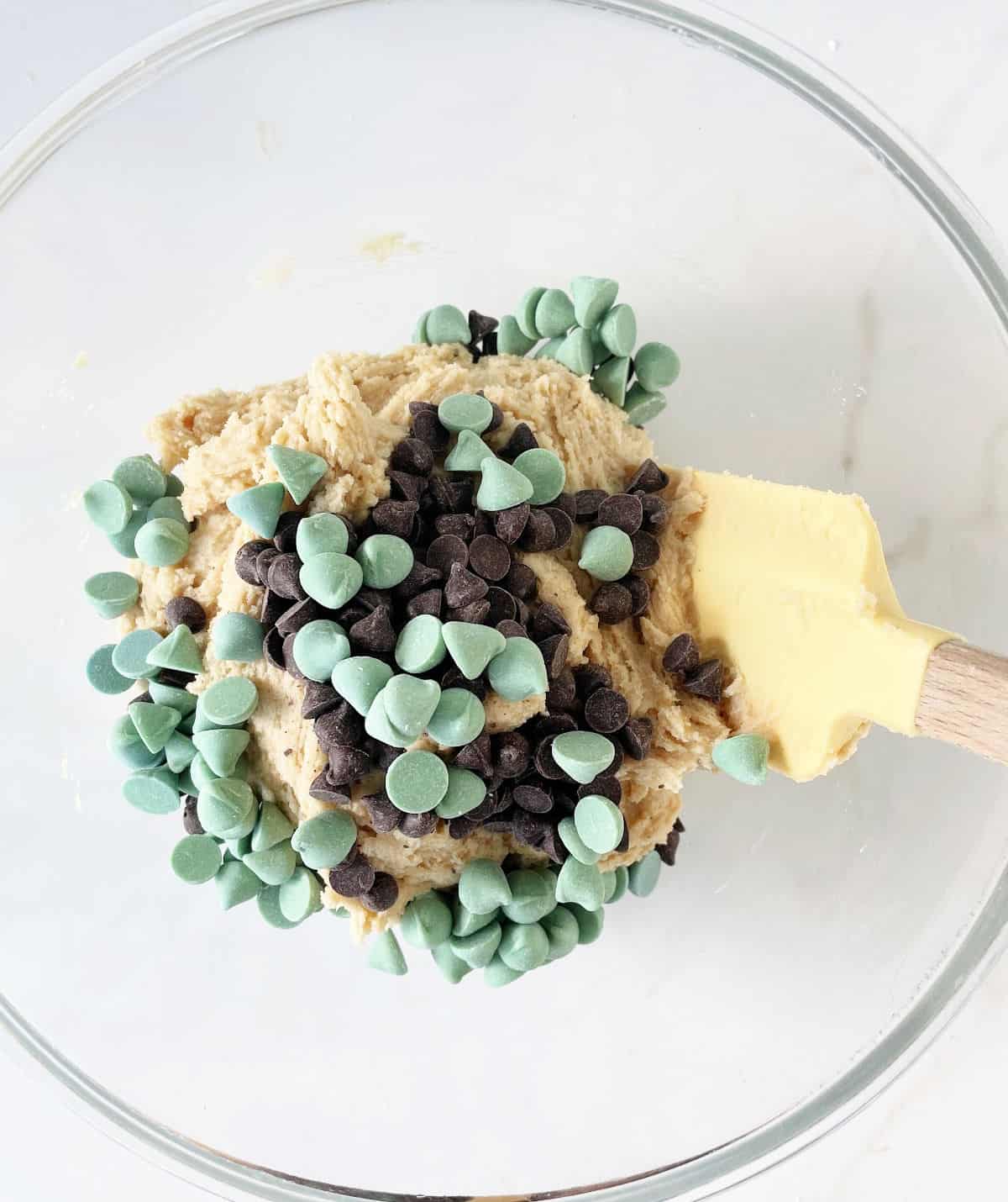 Chocolate and green mint chips added to cookie dough in a glass bowl on a white surface with a yellow spatula inside.